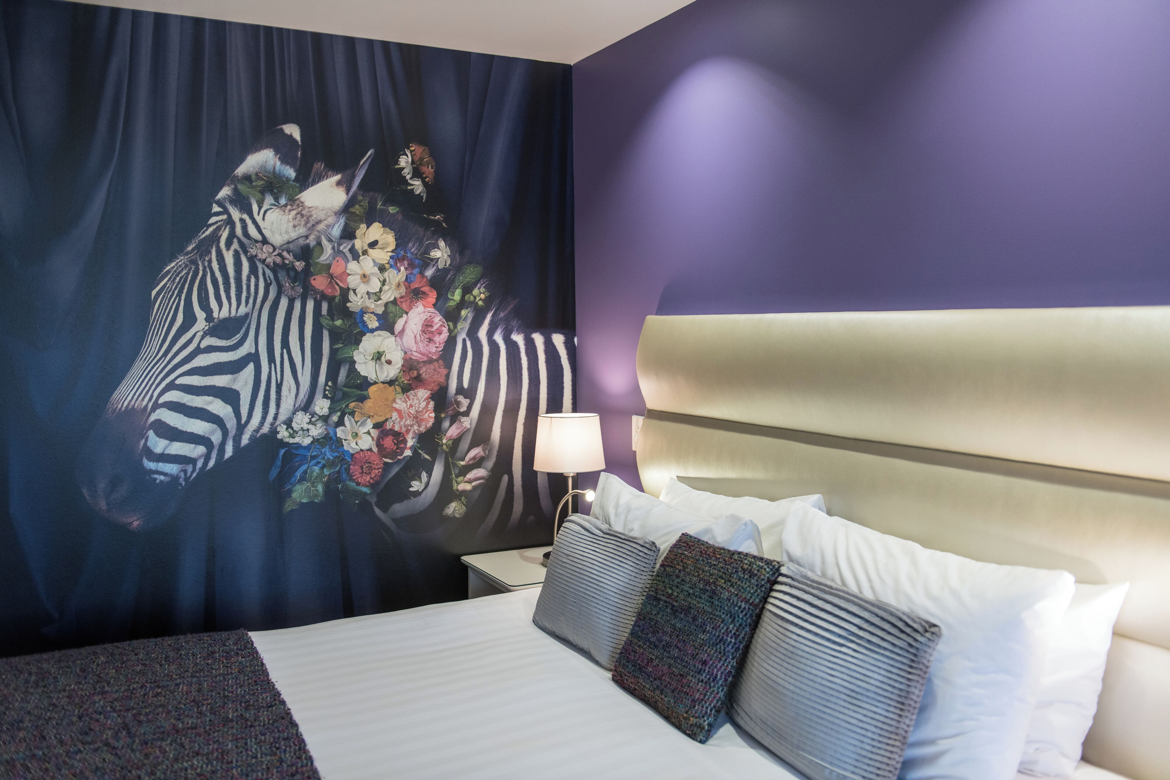 Habitación de hotel con un mural de cebra, una cama con ropa blanca, cabecero y mesita de noche con lámpara.