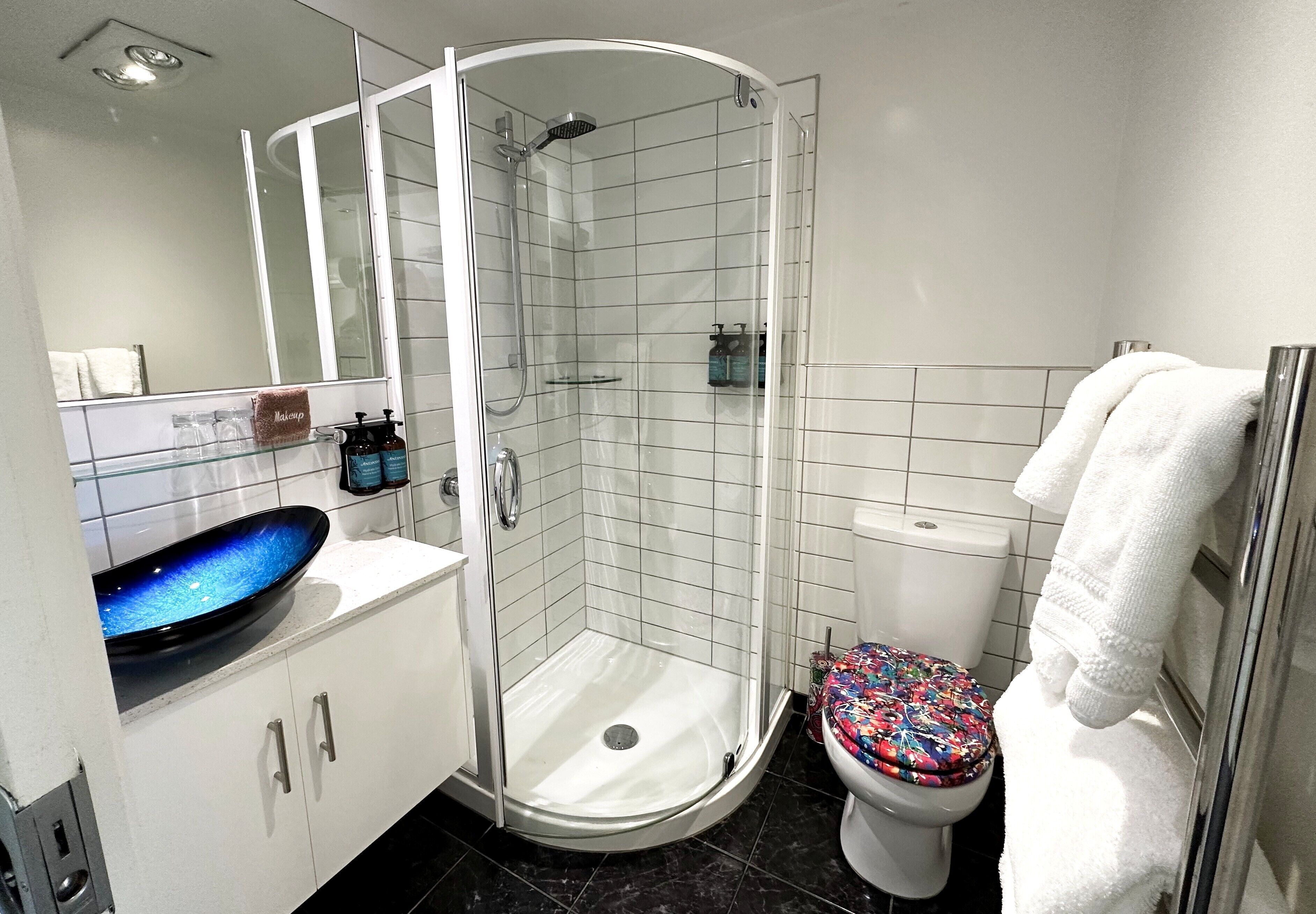 Une salle de bain moderne avec une douche à parois de verre, des toilettes blanches dotées d’un abattant coloré et un lavabo avec un récipient en verre bleu.