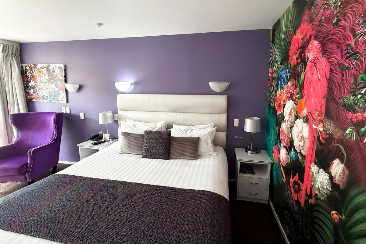 A hotel room with a large bed, a purple armchair, a nightstand with a lamp, and a wall mural of birds and flowers.