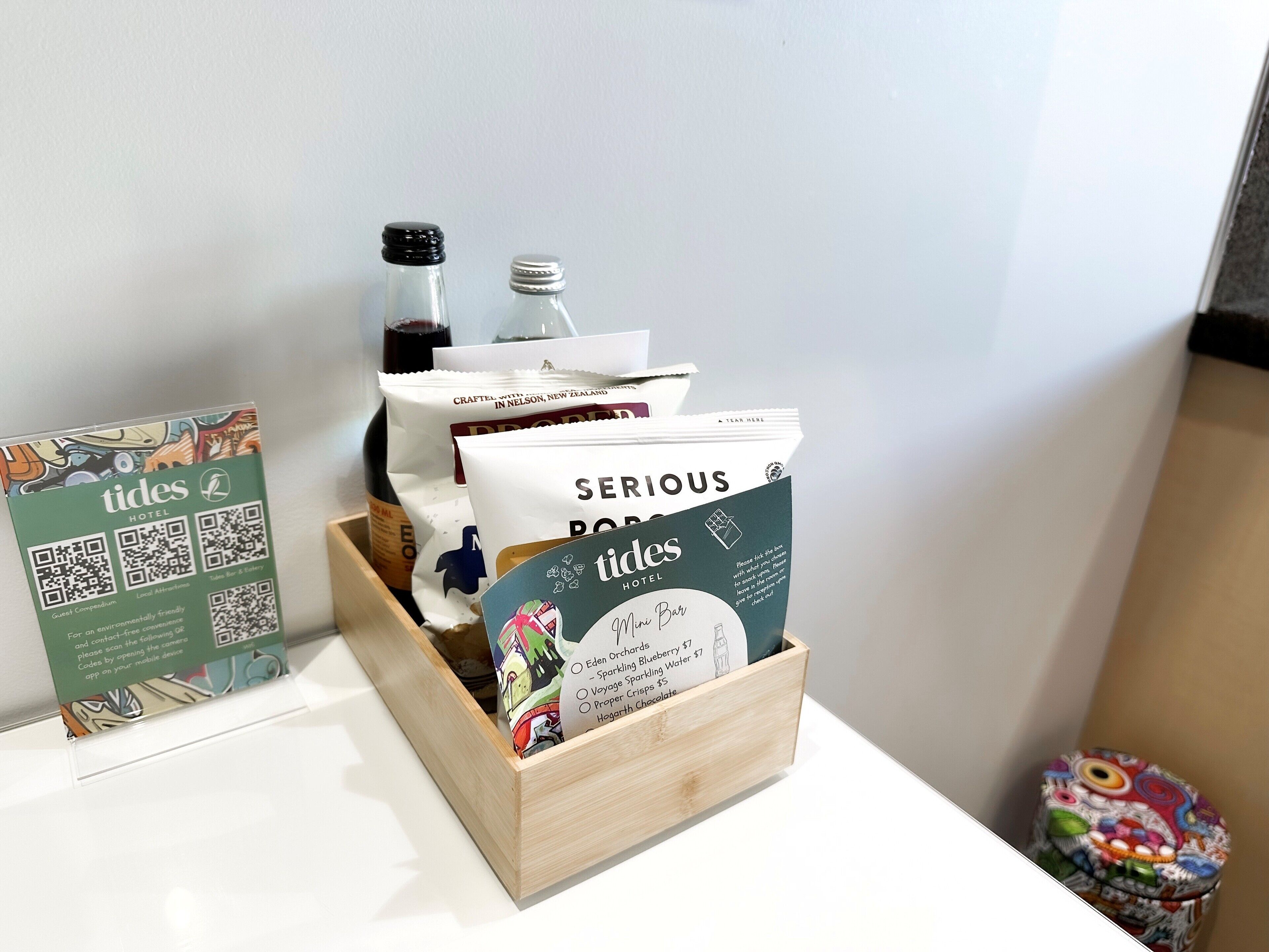 Una caja de madera que contiene varios objetos, incluyendo una botella, una bolsa y una tarjeta con la inscripción 'SERIOUS ROSES' y 'tides HOTEL'.