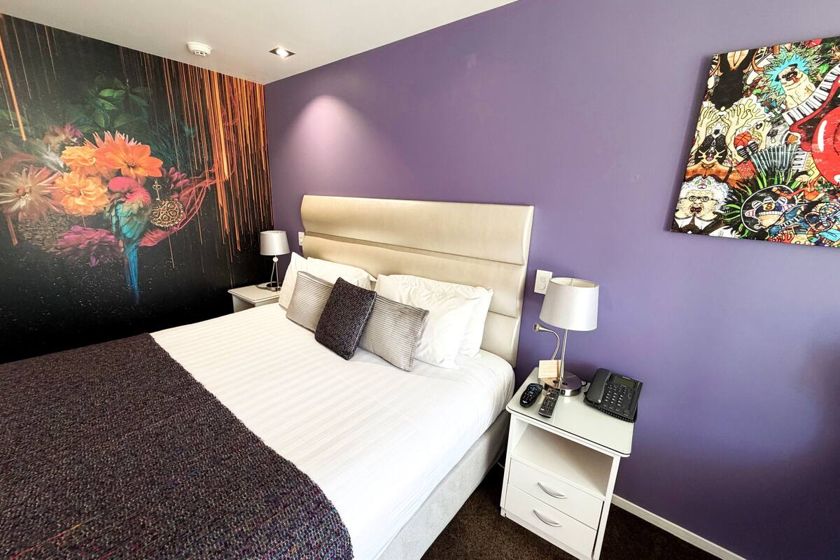 A hotel room with a large bed, a nightstand with a lamp, a phone, and a wall painting.