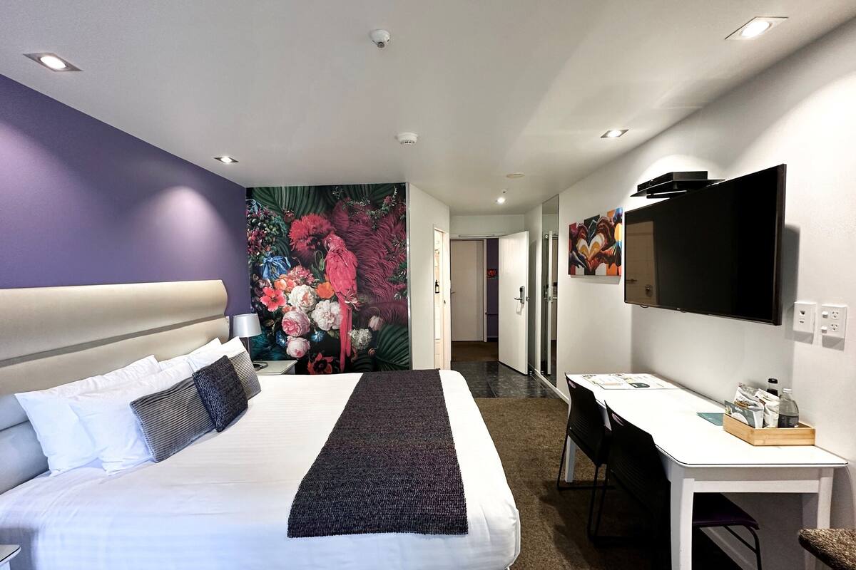 A hotel room with a large bed, a desk, a chair, a TV, and a wall with a floral mural.