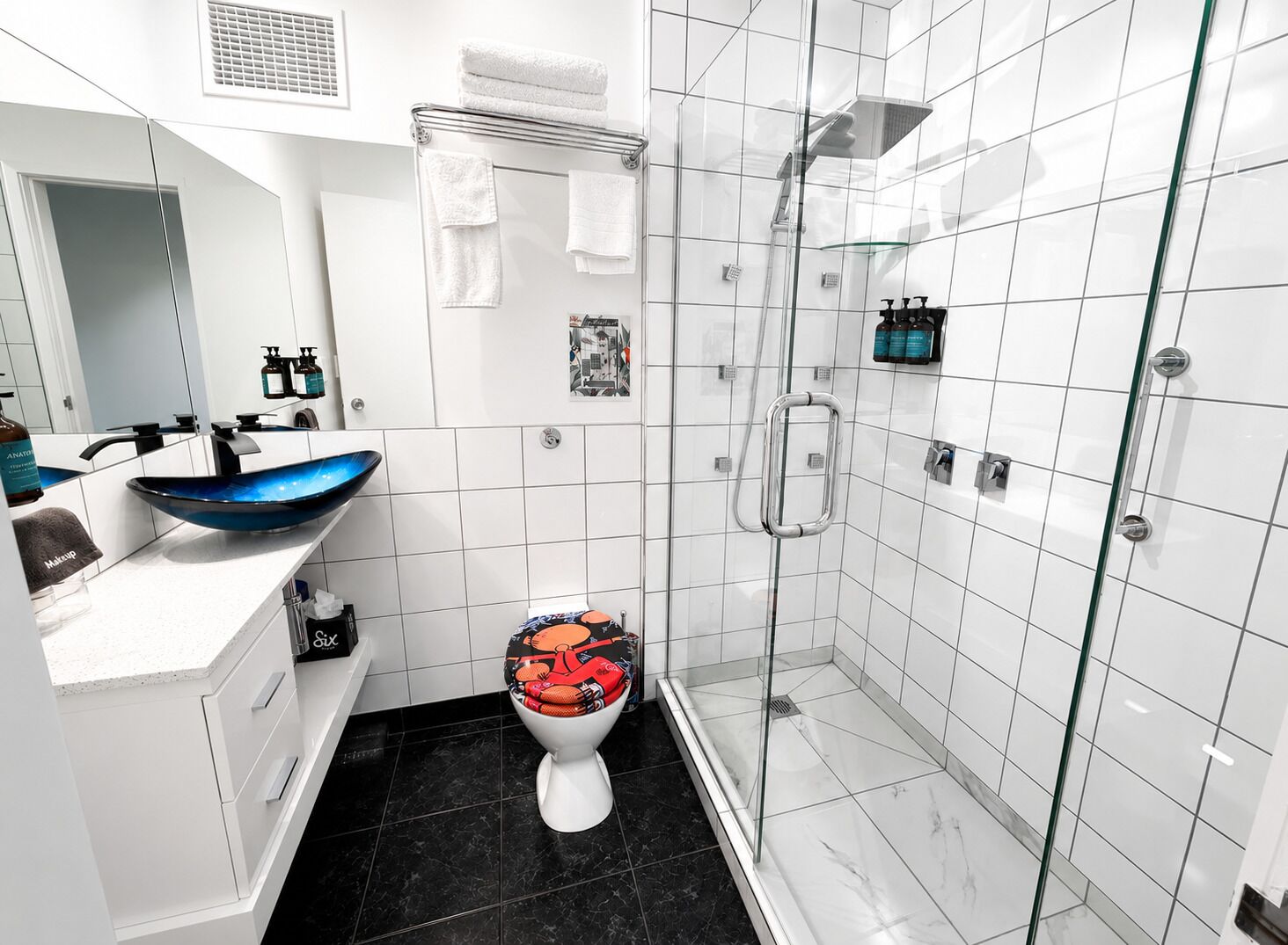 Un baño moderno con ducha de mampara de vidrio, lavamanos blanco con recipiente azul y un inodoro con asiento decorativo.