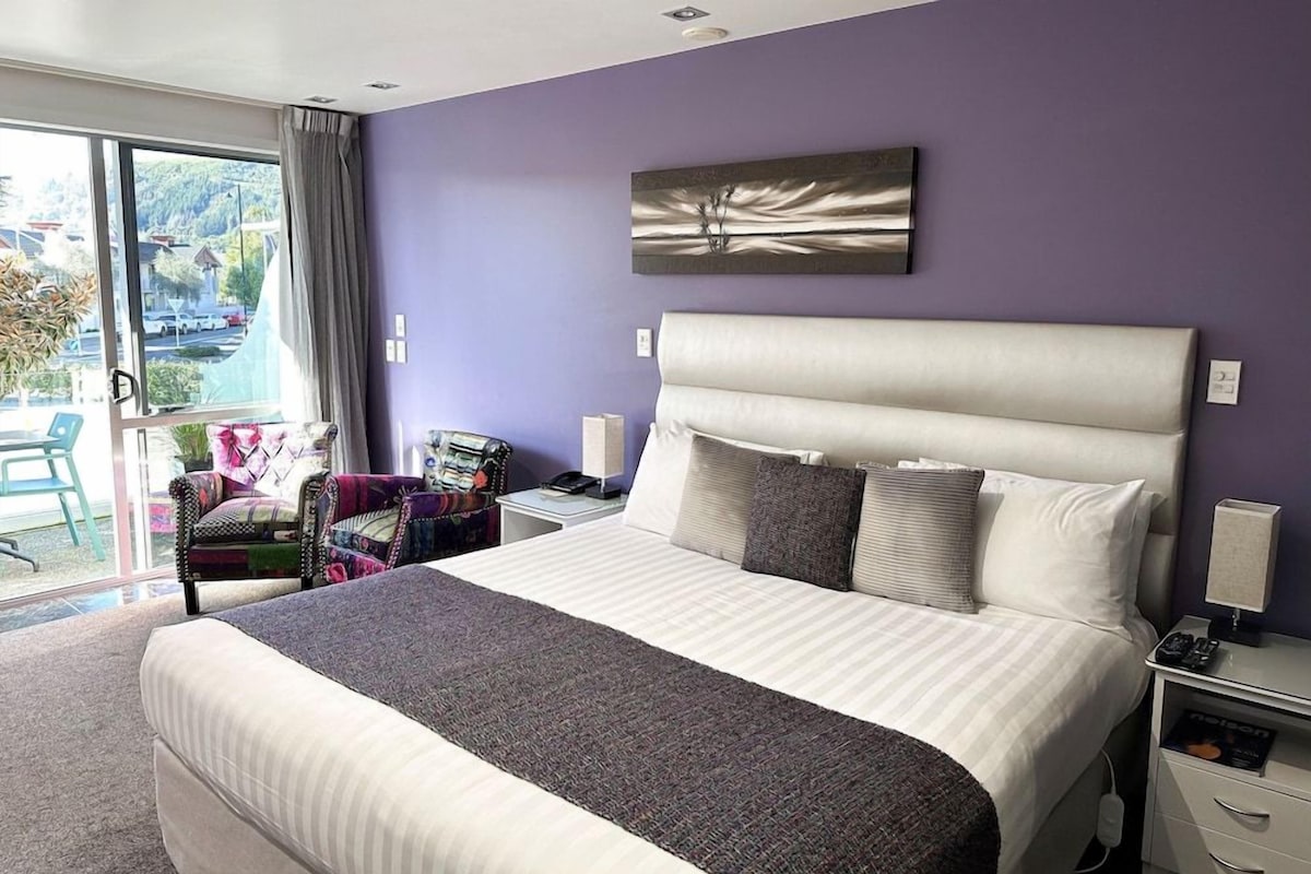 Quarto de hotel com uma cama grande, mesa de cabeceira, cadeira e vista para o exterior.