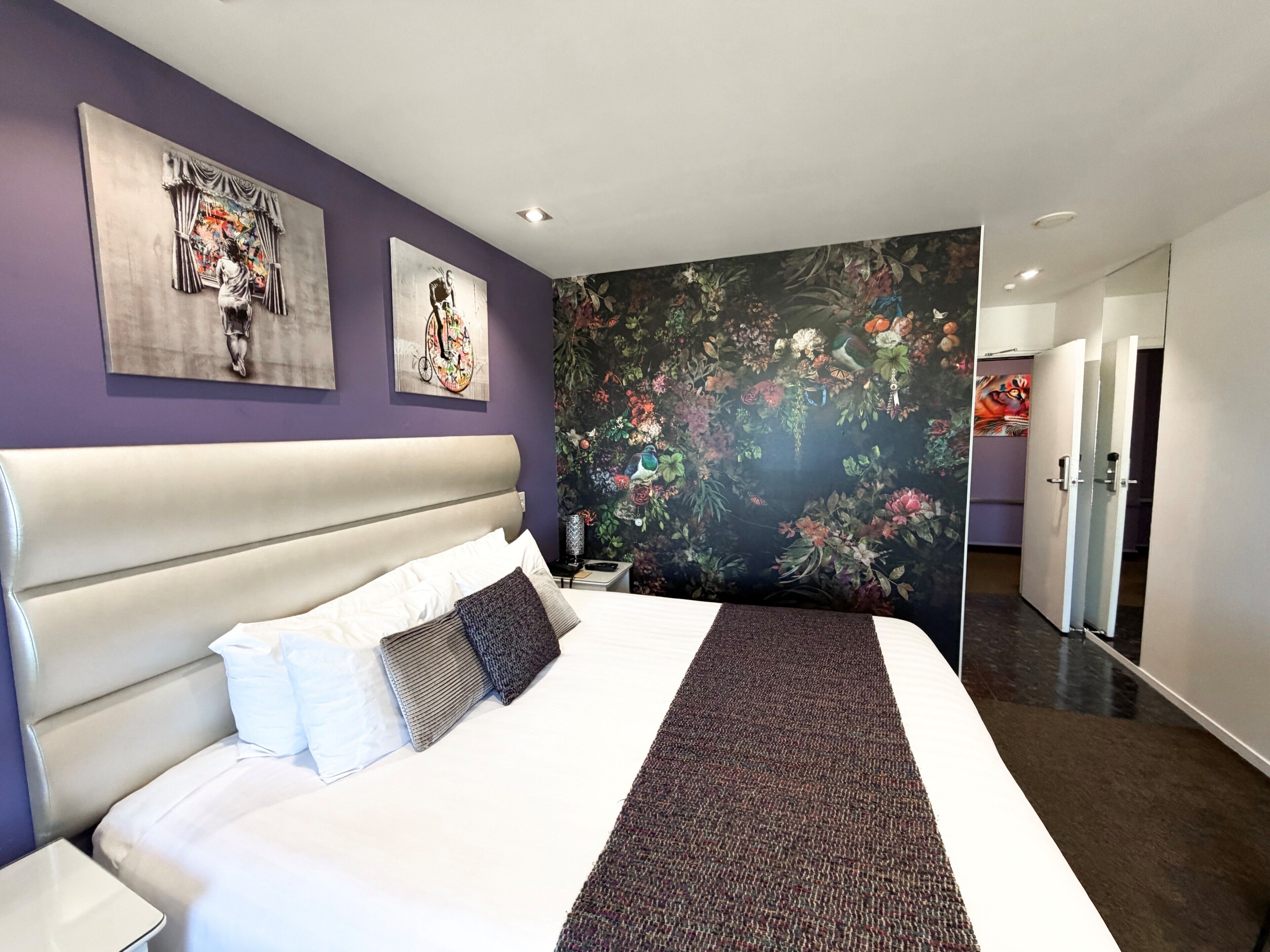 A modern hotel room with a large bed, a headboard, a nightstand, and a floral wallpaper.