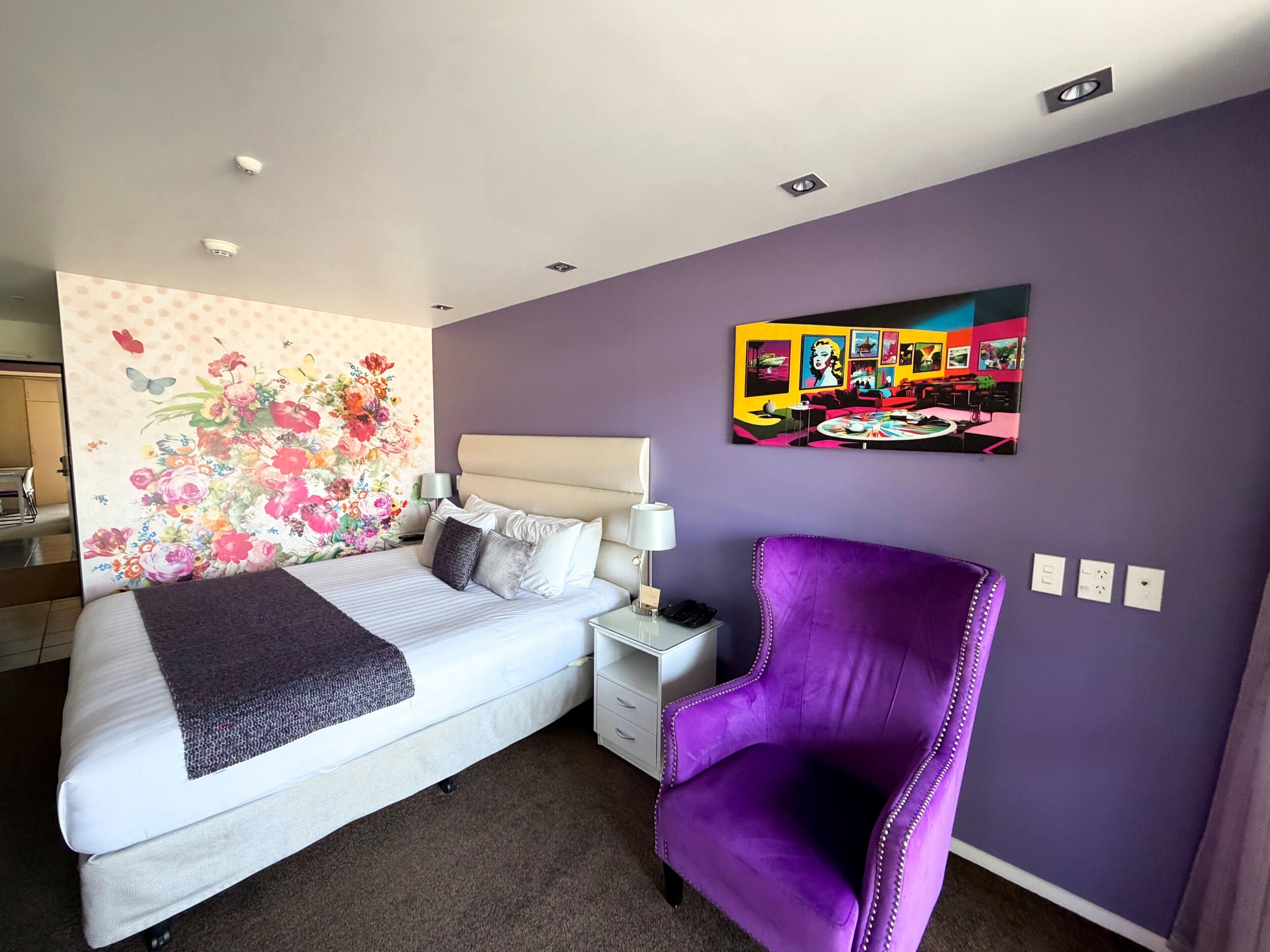 Ein Hotelzimmer mit einem Bett, einem violetten Sessel, einer blumigen Wanddekoration und einem farbenfrohen Kunstwerk an der Wand.