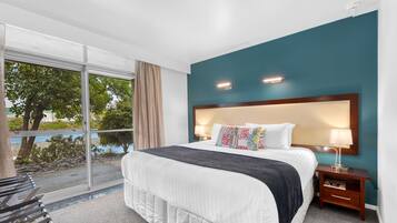 Een hotelkamer met een groot bed, een nachtkastje, een raam met uitzicht op bomen en water, en een balkon met een stoel.