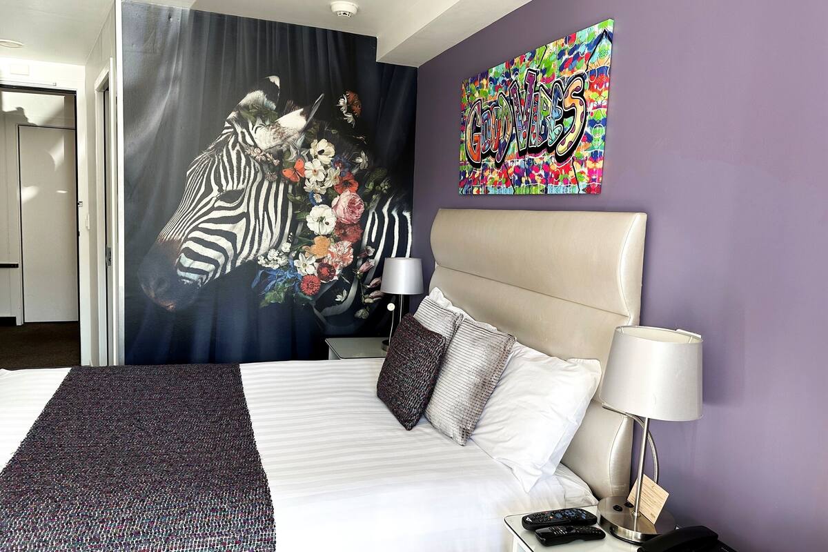 A hotel room with a bed, a headboard, a nightstand with a lamp, a remote control, and a phone.