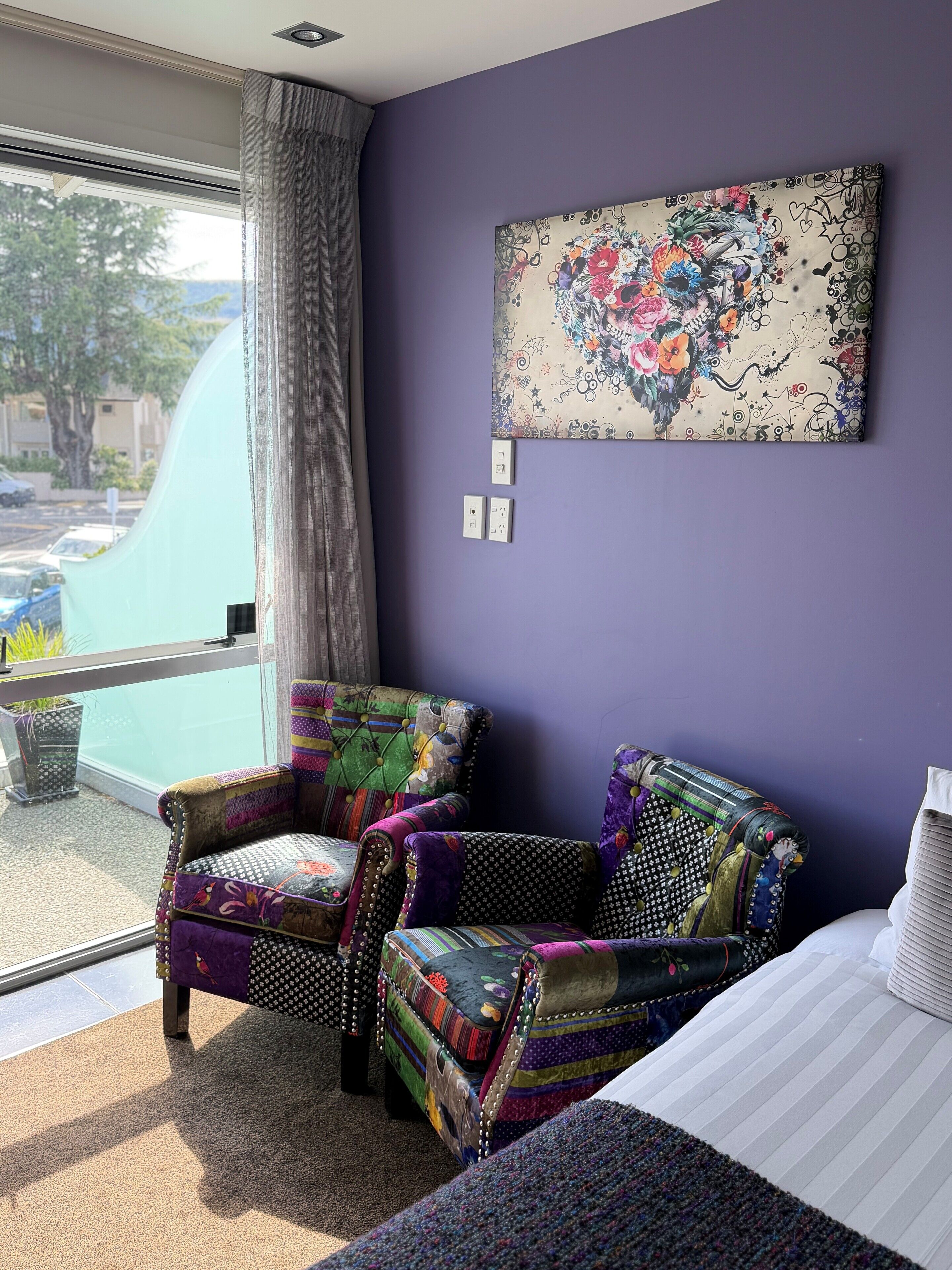 A room with a purple wall, a large window with curtains, a colorful chair, a bed with striped bedding, and a wall-mounted artwork.
