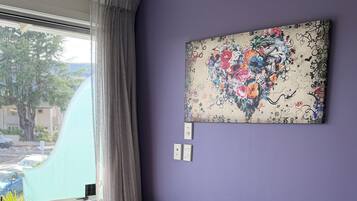 A room with a purple wall, a large window with curtains, a colorful chair, a bed with striped bedding, and a wall-mounted artwork.