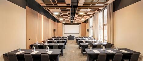 A conference room with multiple tables and chairs, a projector screen at the front, and large windows on one side.