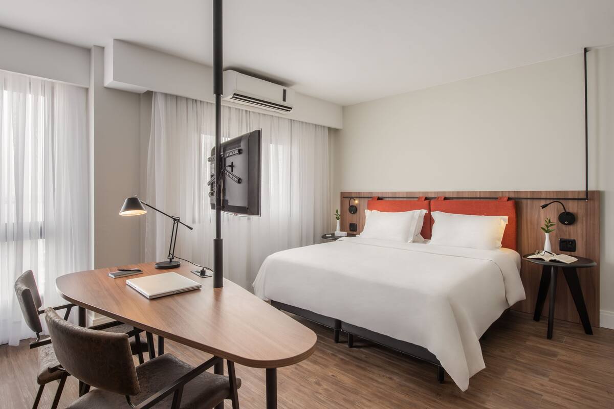 Een hotelkamer met een houten bureau, een bed met wit beddengoed, een nachtkastje en een aan de muur bevestigde televisie.