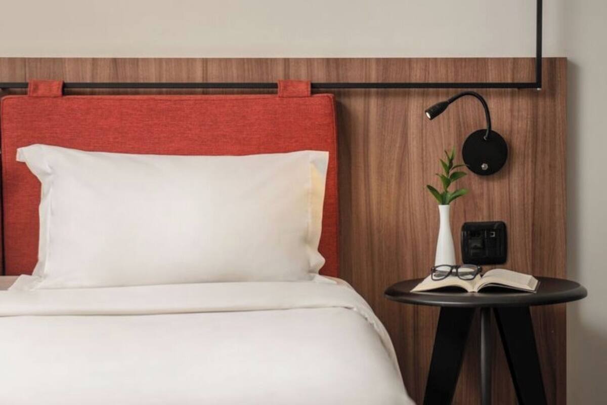 Een netjes opgemaakt bed met een rode hoofdeinde, een klein rond tafeltje met een plant, glazen en een open boek, en een wandlamp.