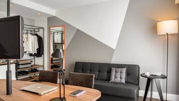 A modern hotel room with a wooden desk, a laptop, a mirror, a black sofa, and a small table with a lamp.