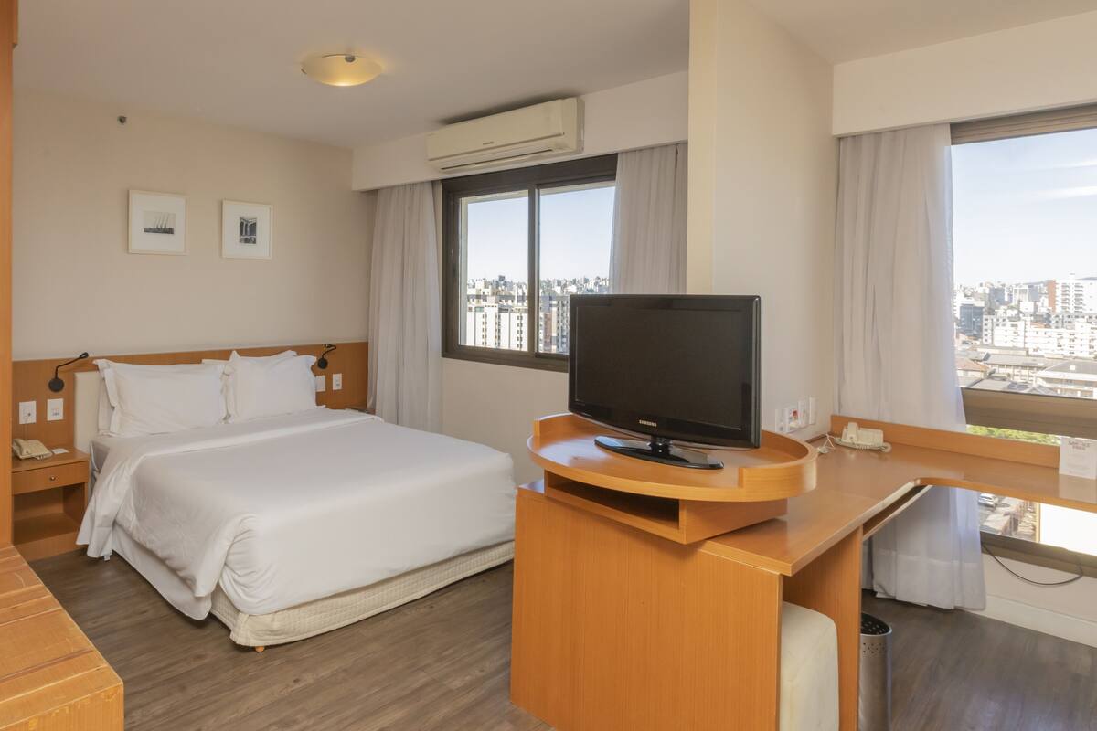 Hotelkamer met een bed, een bureau met televisie, een raam met gordijnen en uitzicht op de stad.