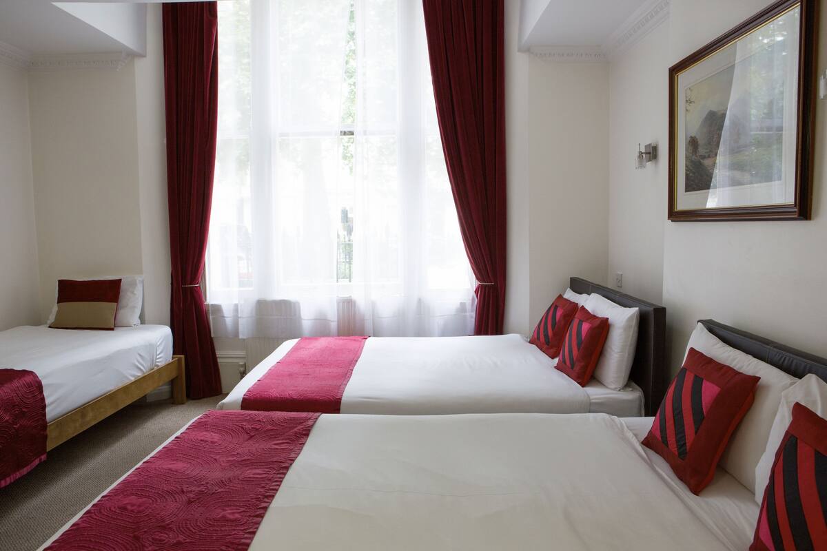 Hotellihuone, jossa on kolme sänkyä, punaiset verhot ja seinällä oleva kehystetty taulu.