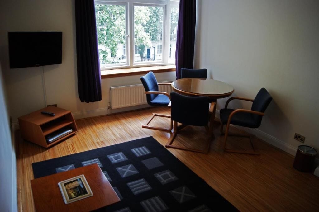 Ein Zimmer mit einem runden Tisch, zwei Stühlen, einem Fernseher, einer Heizung und einem Fenster mit Vorhängen.