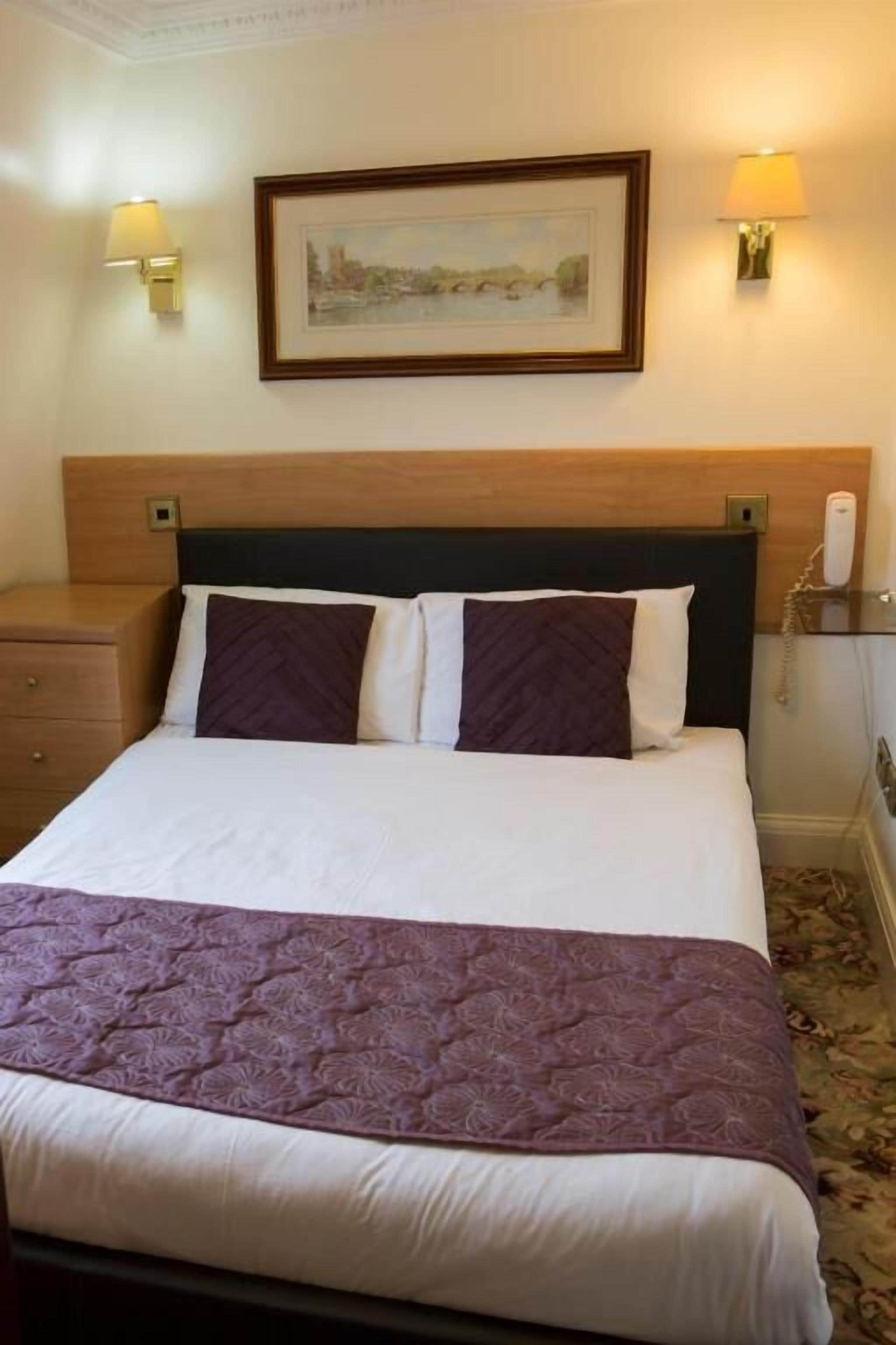 A hotel room with a bed, two bedside lamps, a framed picture on the wall, and a telephone on the nightstand.