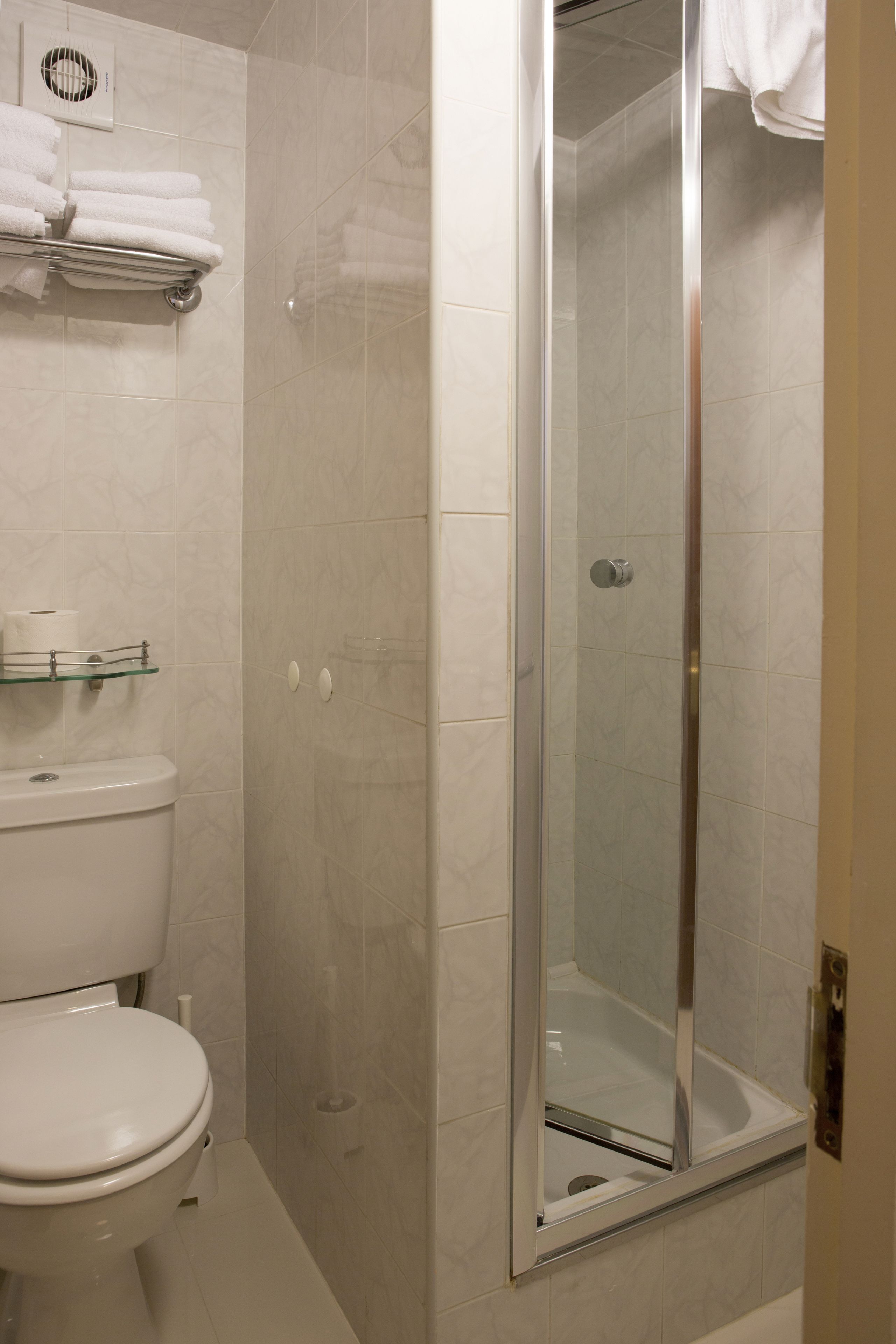 A bathroom with a toilet, a glass shelf with towels, and a glass-enclosed shower area.