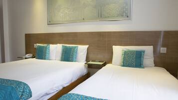 A hotel room with two beds, each with white linens and teal decorative pillows. There is a framed artwork above the beds, a bedside table with a phone, and a nightstand with a lamp.