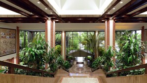 A spacious indoor area with a central staircase, surrounded by lush greenery and large windows offering views of the outdoors.