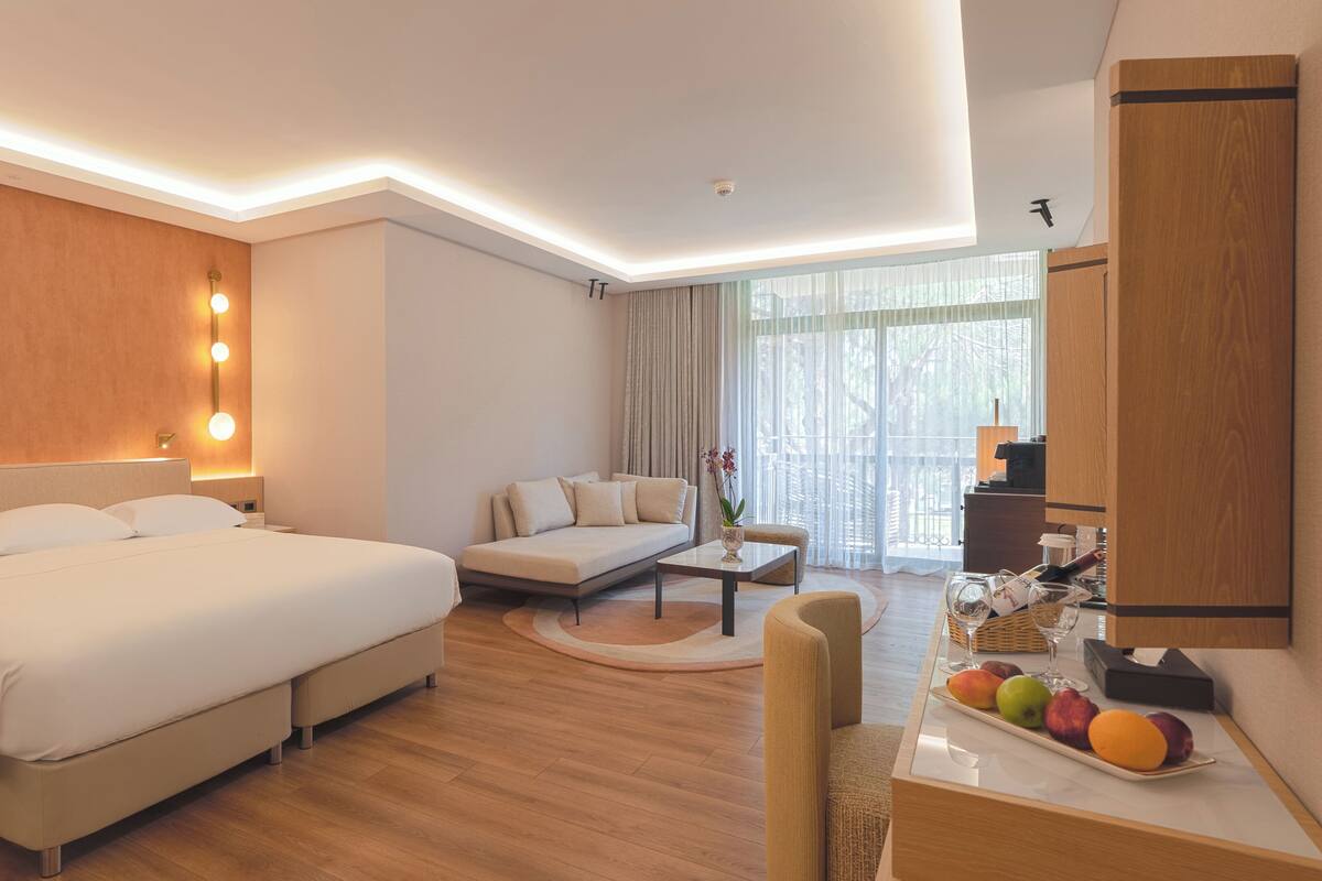 A hotel room with a large bed, a sofa, a dining table with fruit, and a view of the outdoors.