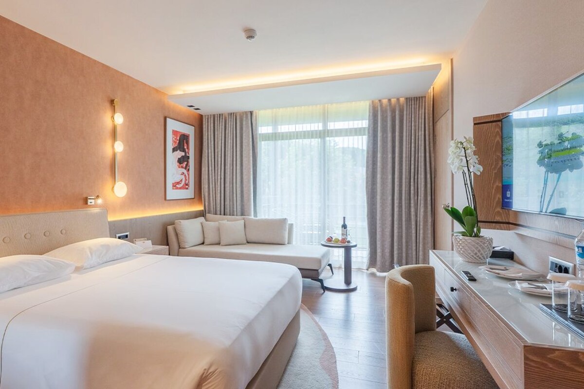 A hotel room with a large bed, a sofa, a desk with a TV, and a small table with a plant.
