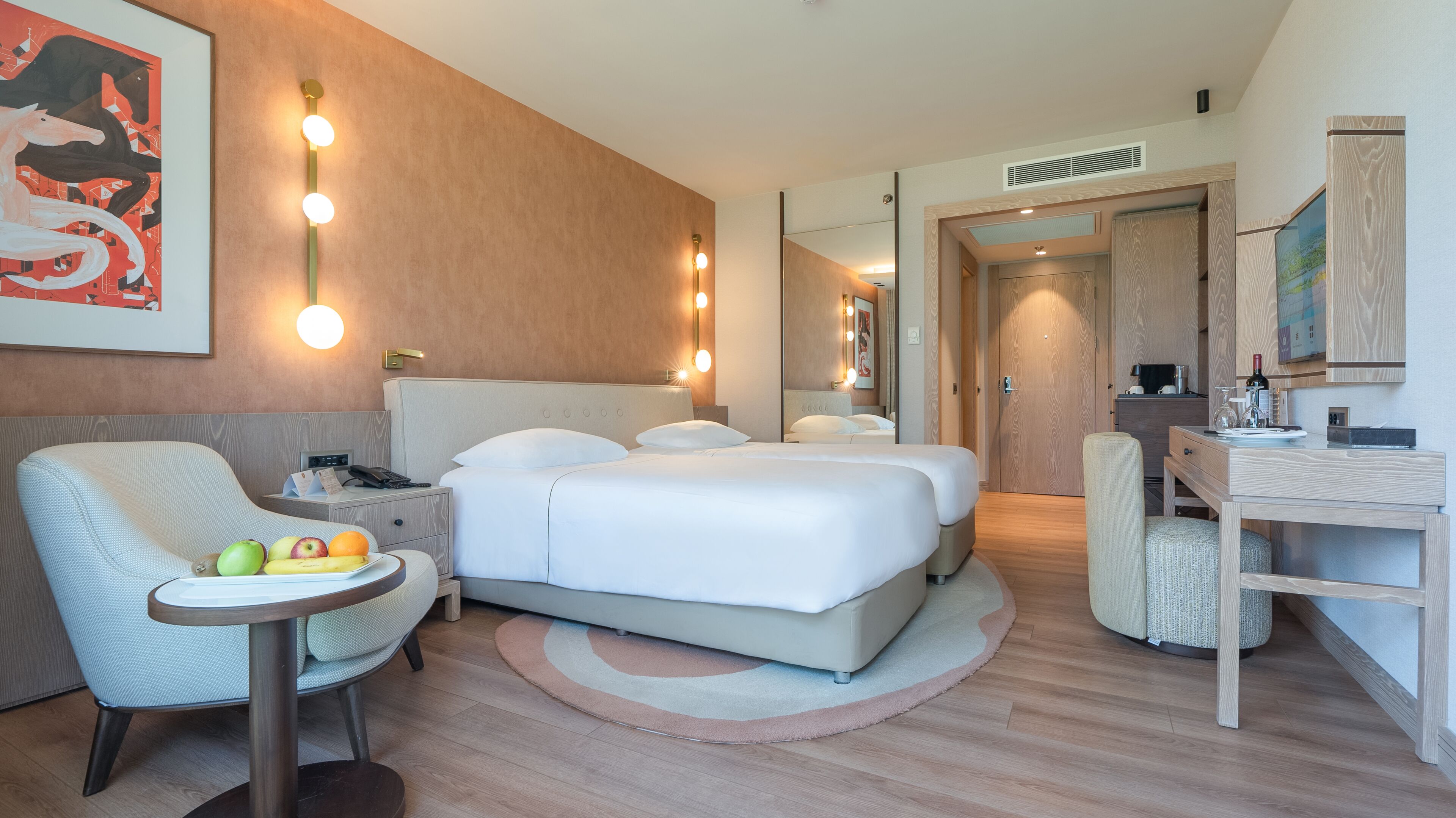 A hotel room with a bed, bedside tables, a chair, a small table with fruit, and a mirror.
