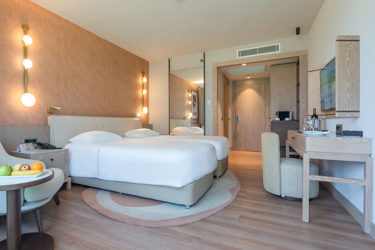 A hotel room with a bed, bedside tables, a chair, a small table with fruit, and a mirror.