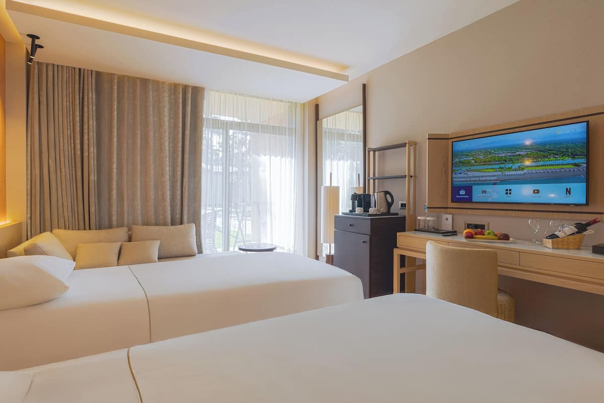 A hotel room with two beds, a desk, a TV, and a view of the outdoors.