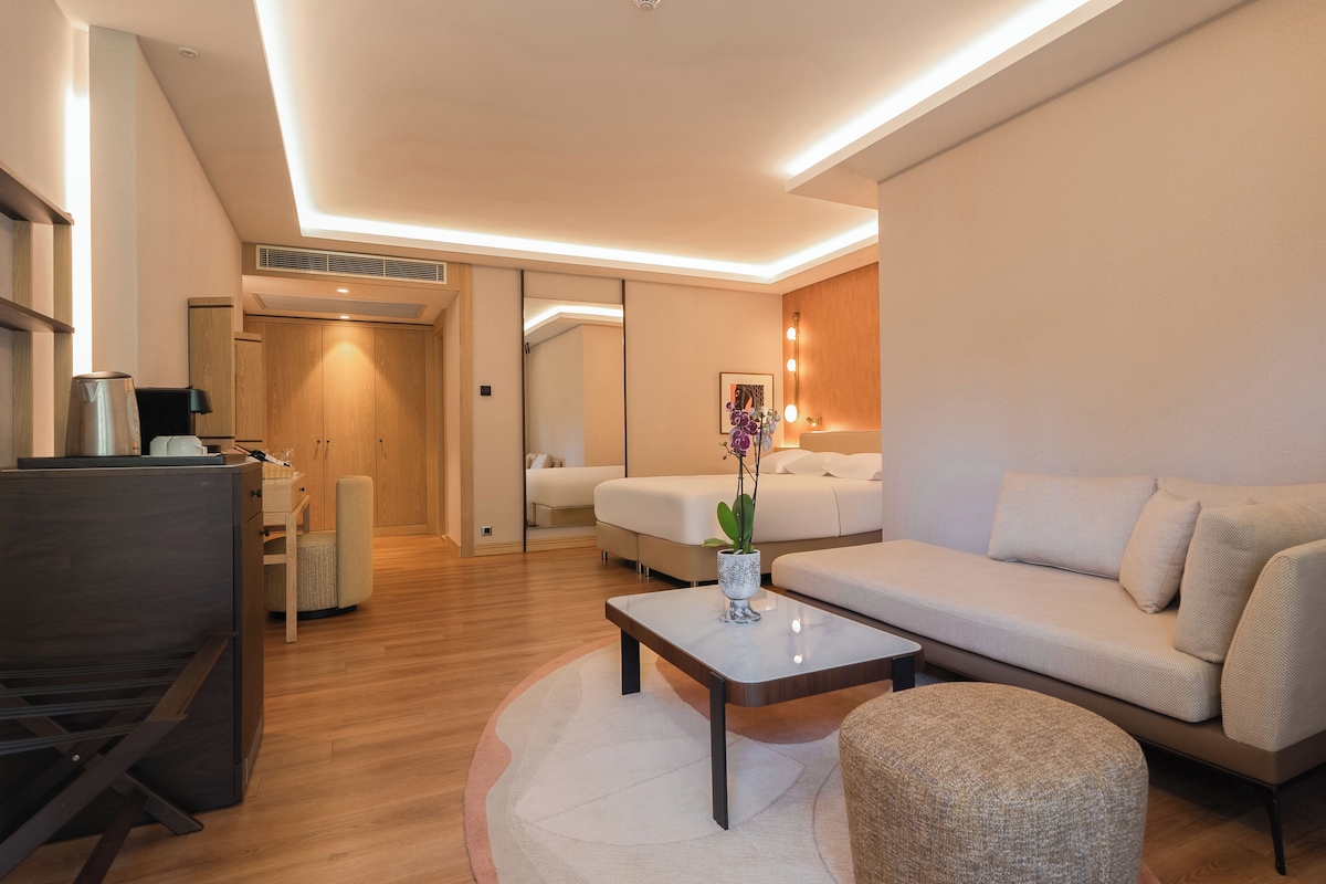 A modern hotel room with a sofa, a coffee table, a small rug, a bedside table, and a lamp.