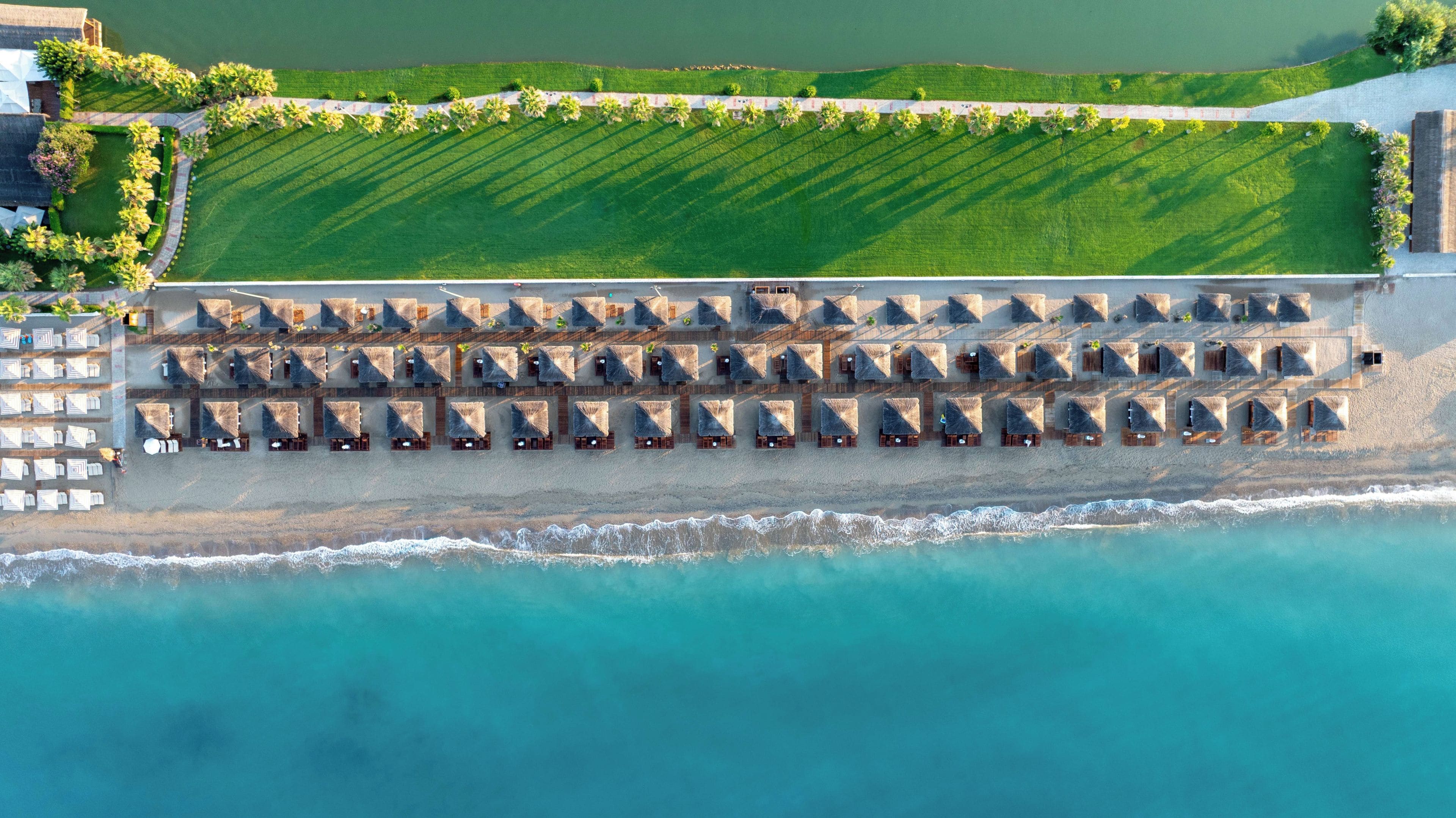 Vue aérienne d’un complexe hôtelier en bord de mer, avec des rangées de cabanes de plage aux toits de chaume et des chaises longues.