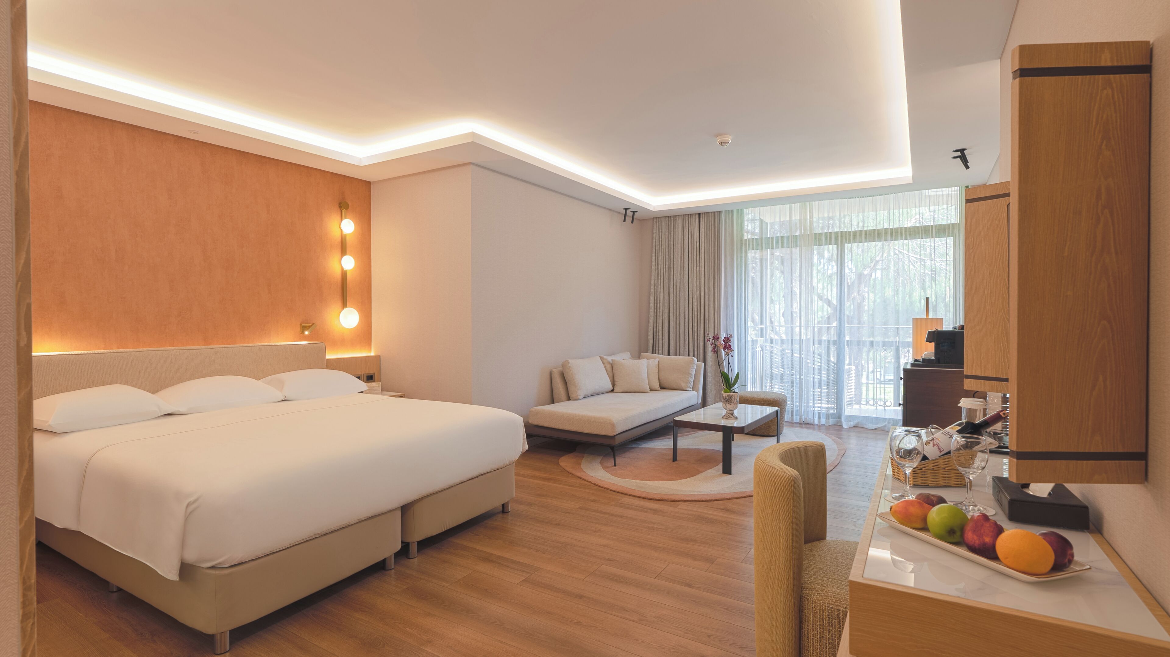 A hotel room with a large bed, a sofa, a dining table with fruit, and a view of the outdoors.