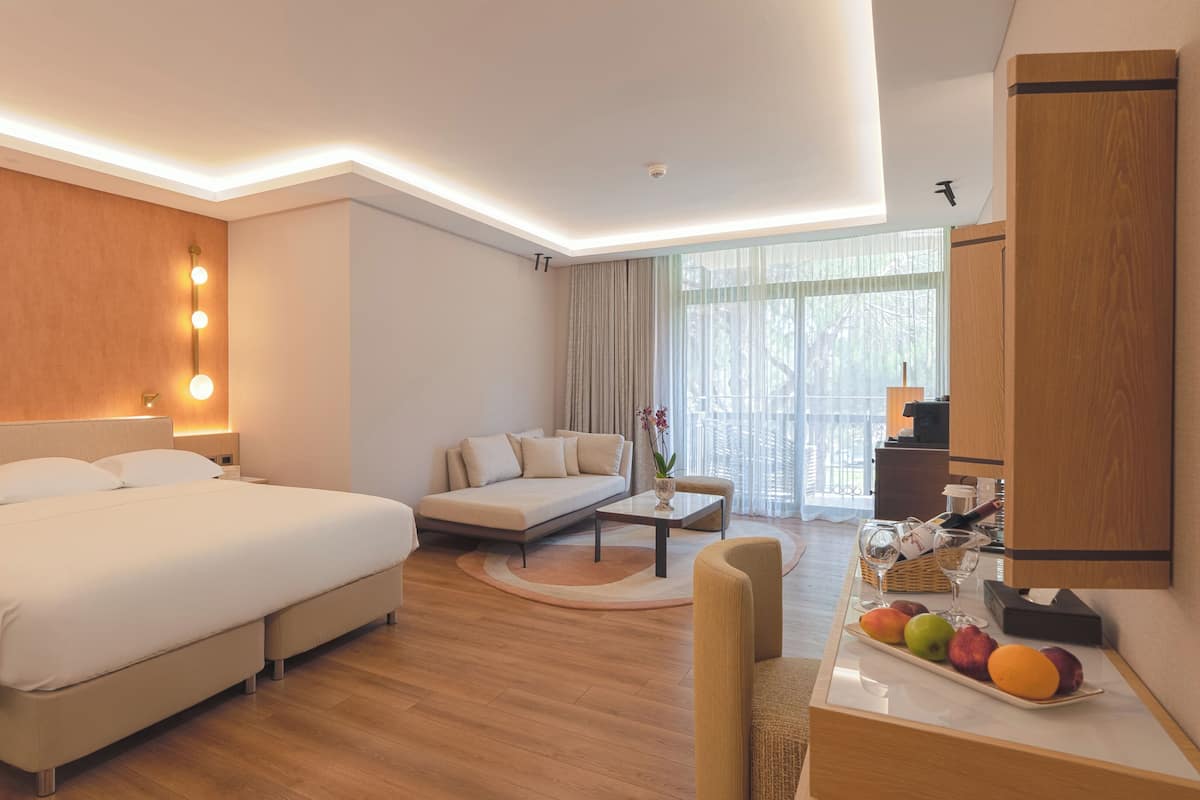 A hotel room with a large bed, a sofa, a dining table with fruit, and a view of the outdoors.