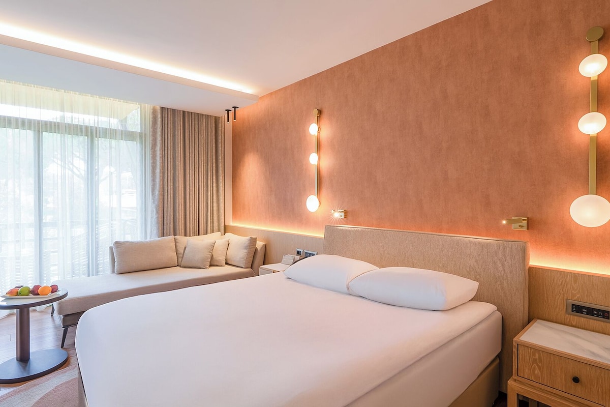 A hotel room with a large bed, a bedside table with fruit, a sofa, and a wall-mounted light fixture.