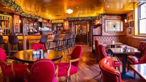 A bar area with wooden flooring, red upholstered chairs, and a bar counter with stools. - Benners Hotel (Tralee)