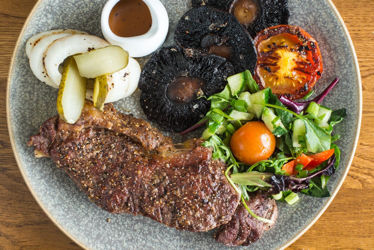 A plate with a steak, pickles, mushrooms, a grilled tomato, and a salad.