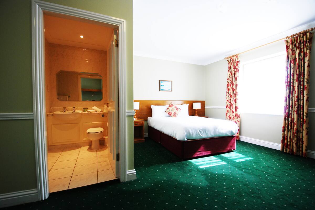 Una habitación de hotel con alfombra verde, una cama con ropa de cama blanca, cabecera de madera, una mesita de noche, y un baño con inodoro, lavabo y espejo.