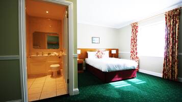 Une chambre d’hôtel avec une moquette verte, un lit avec une literie blanche, une tête de lit en bois, une table de chevet et une salle de bain comprenant des toilettes, un lavabo et un miroir.