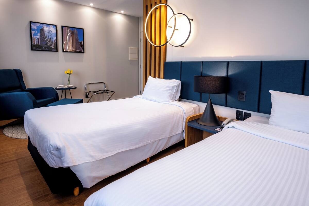 Ein Hotelzimmer mit zwei Betten, einem Schreibtisch, einem Stuhl, einer Lampe und gerahmten Bildern an der Wand.