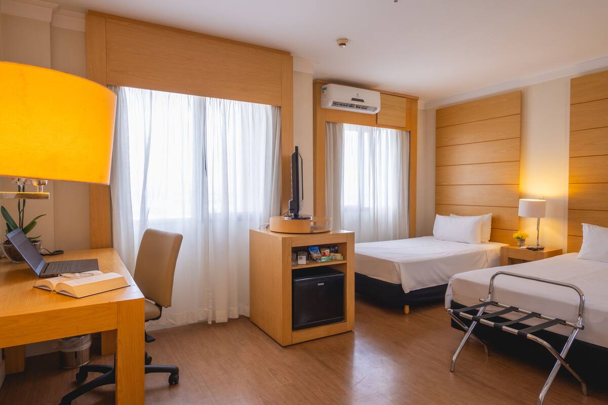 Ein Hotelzimmer mit zwei Betten, einem Schreibtisch mit Laptop, einem kleinen Kühlschrank, einem Fernseher und einer Lampe.
