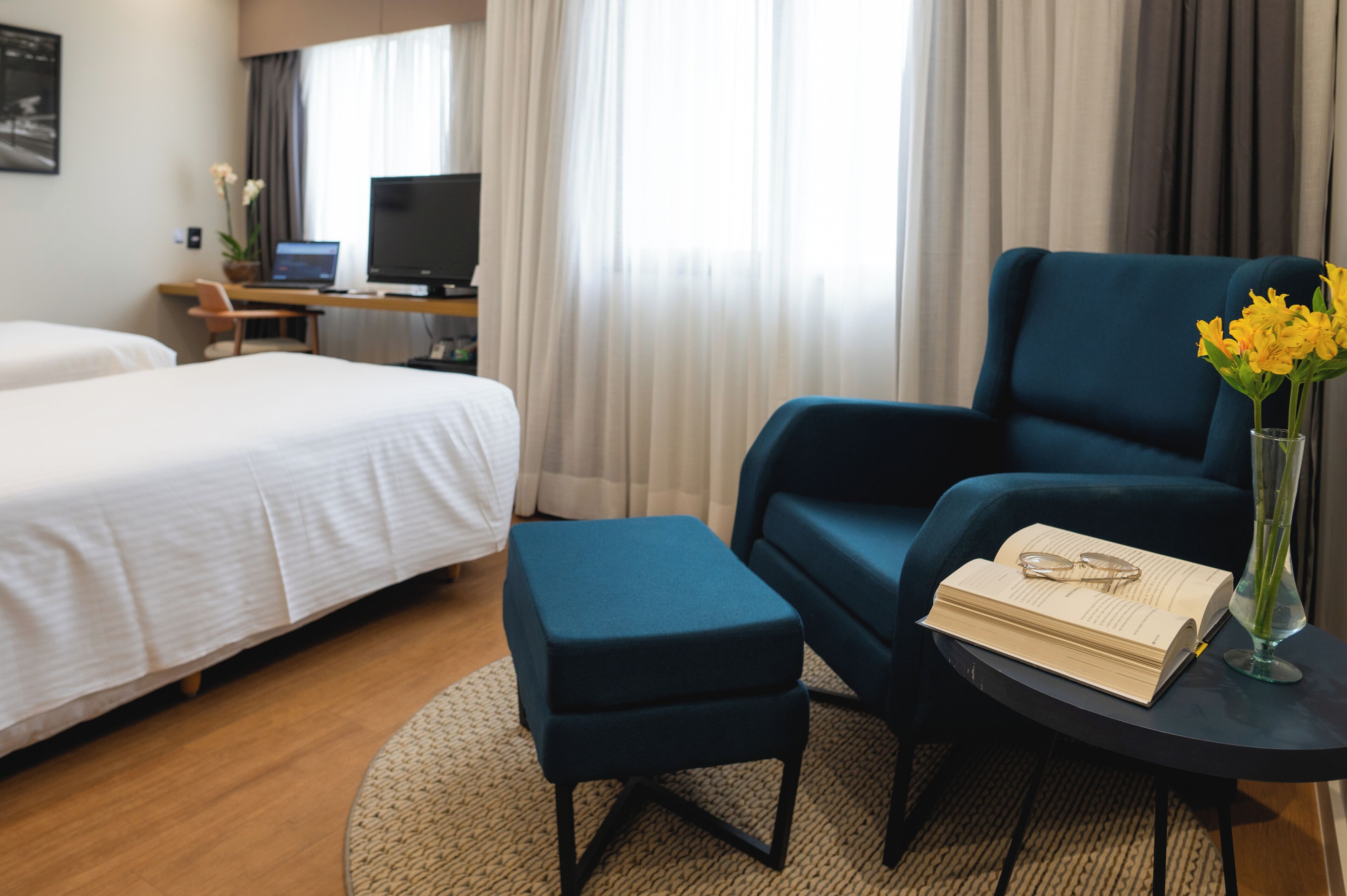 Habitación de hotel con dos camas, un escritorio con computadora, un sillón azul y una mesita con un libro y gafas.