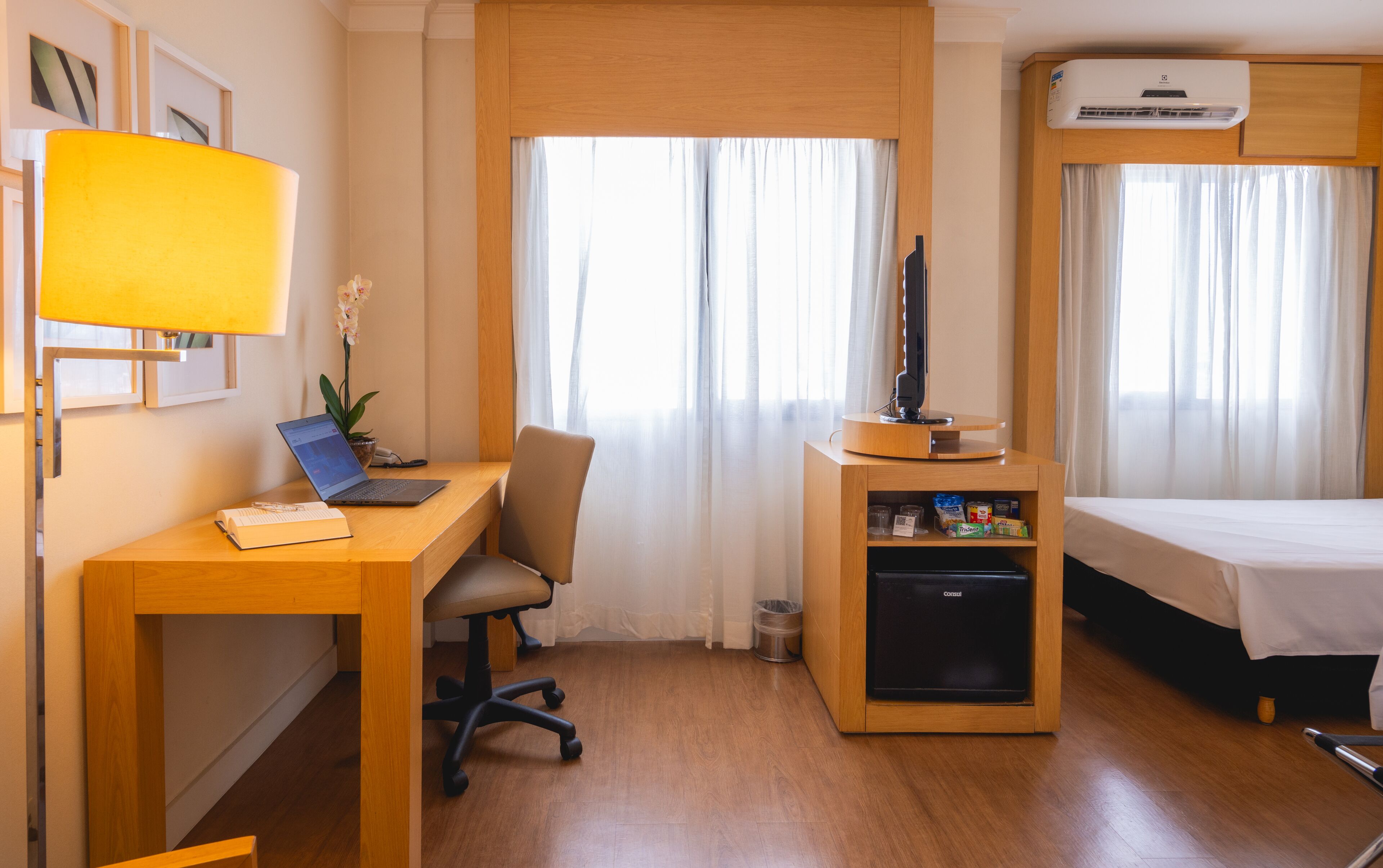 Habitación de hotel con escritorio de madera, una computadora portátil, un televisor pequeño sobre un soporte de madera, un refrigerador minibar, una cama y una ventana con cortinas.