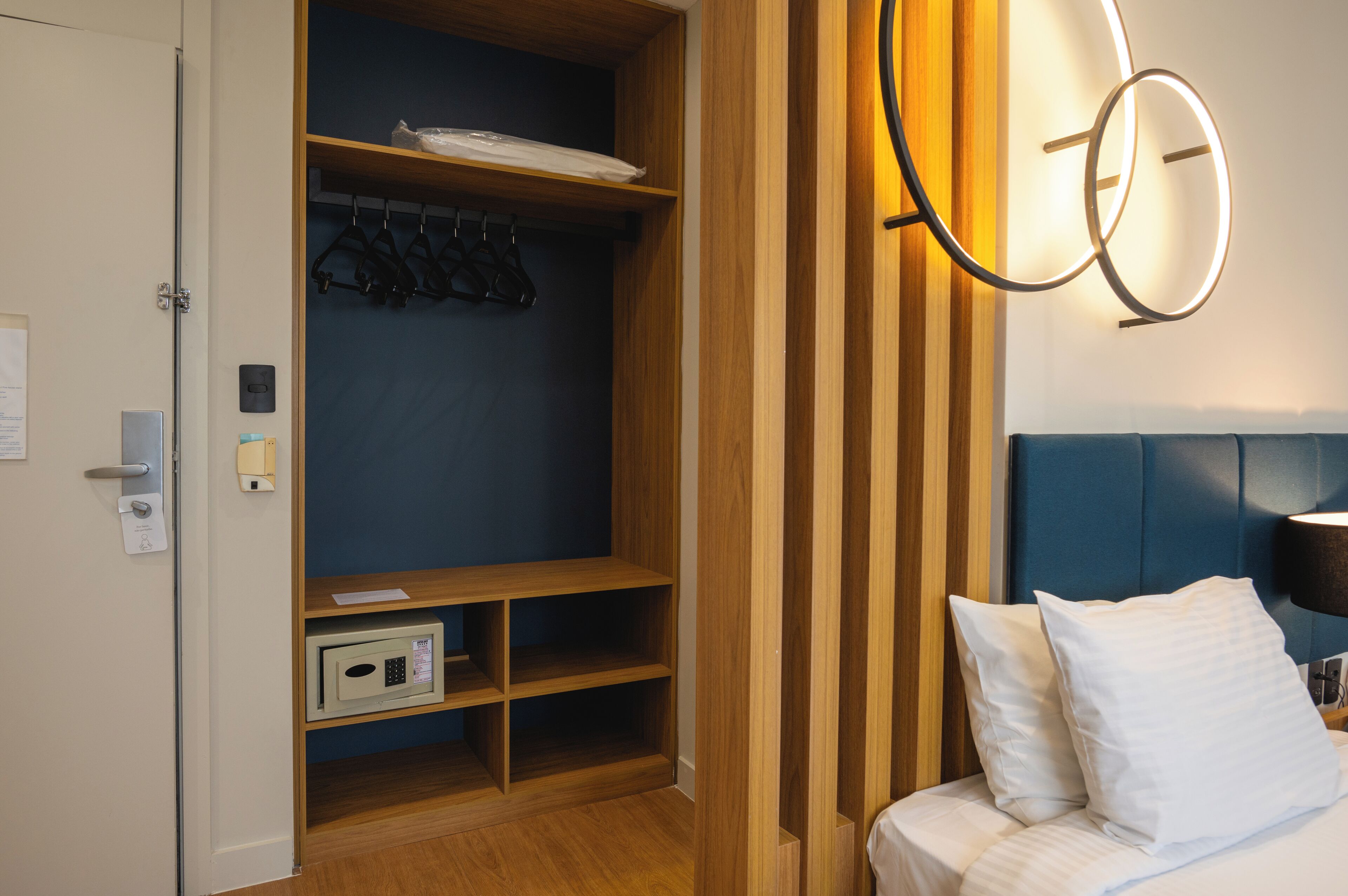 A hotel room with a wardrobe, a bed with white bedding, and a wall-mounted light fixture.