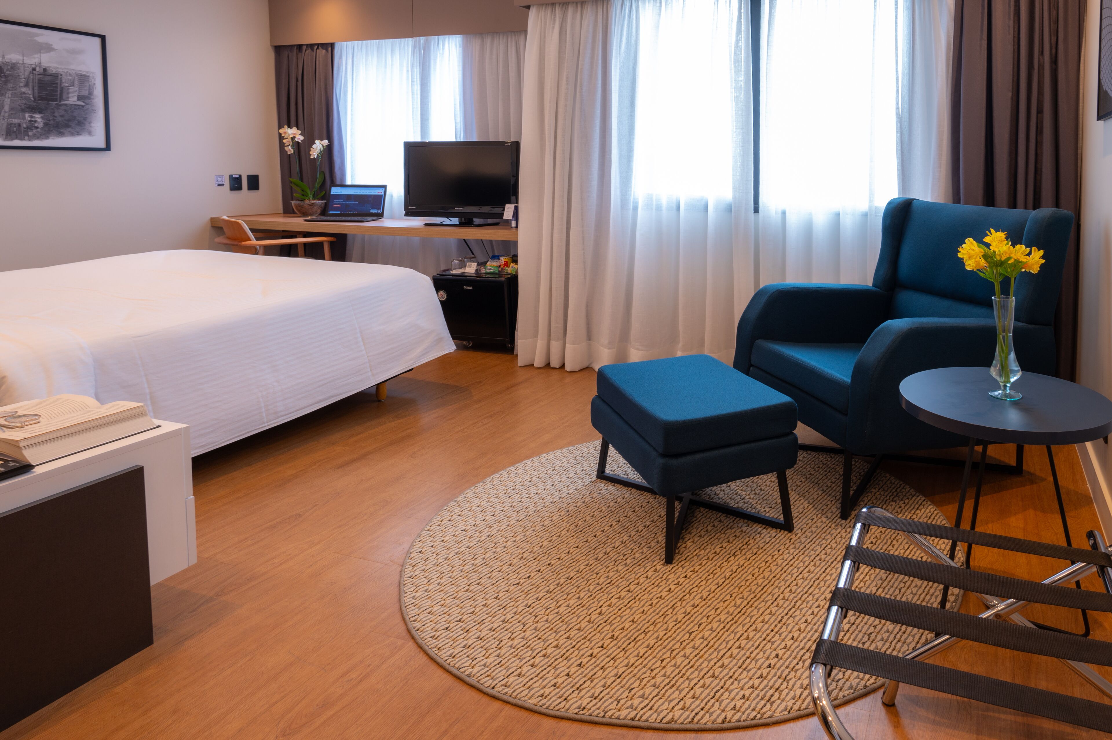 Ett hotellrum med en säng, ett skrivbord med en bärbar dator, en tv, en blå fåtölj, ett litet bord med en vas med blommor och ett fönster med gardiner.
