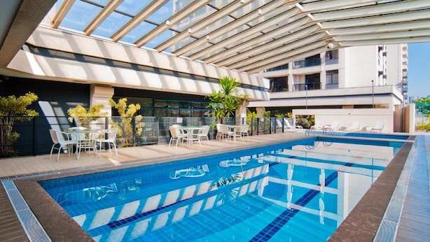 Una piscina interior moderna con agua azul cristalina, rodeada de sillones y mesas, y una estructura de techo de cristal.