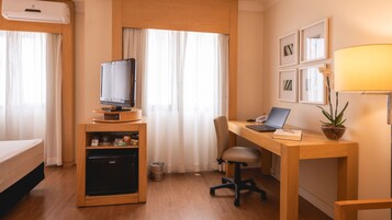 Habitación de hotel con cama, escritorio con computadora portátil, televisor sobre un mueble, refrigerador pequeño y planta en maceta.