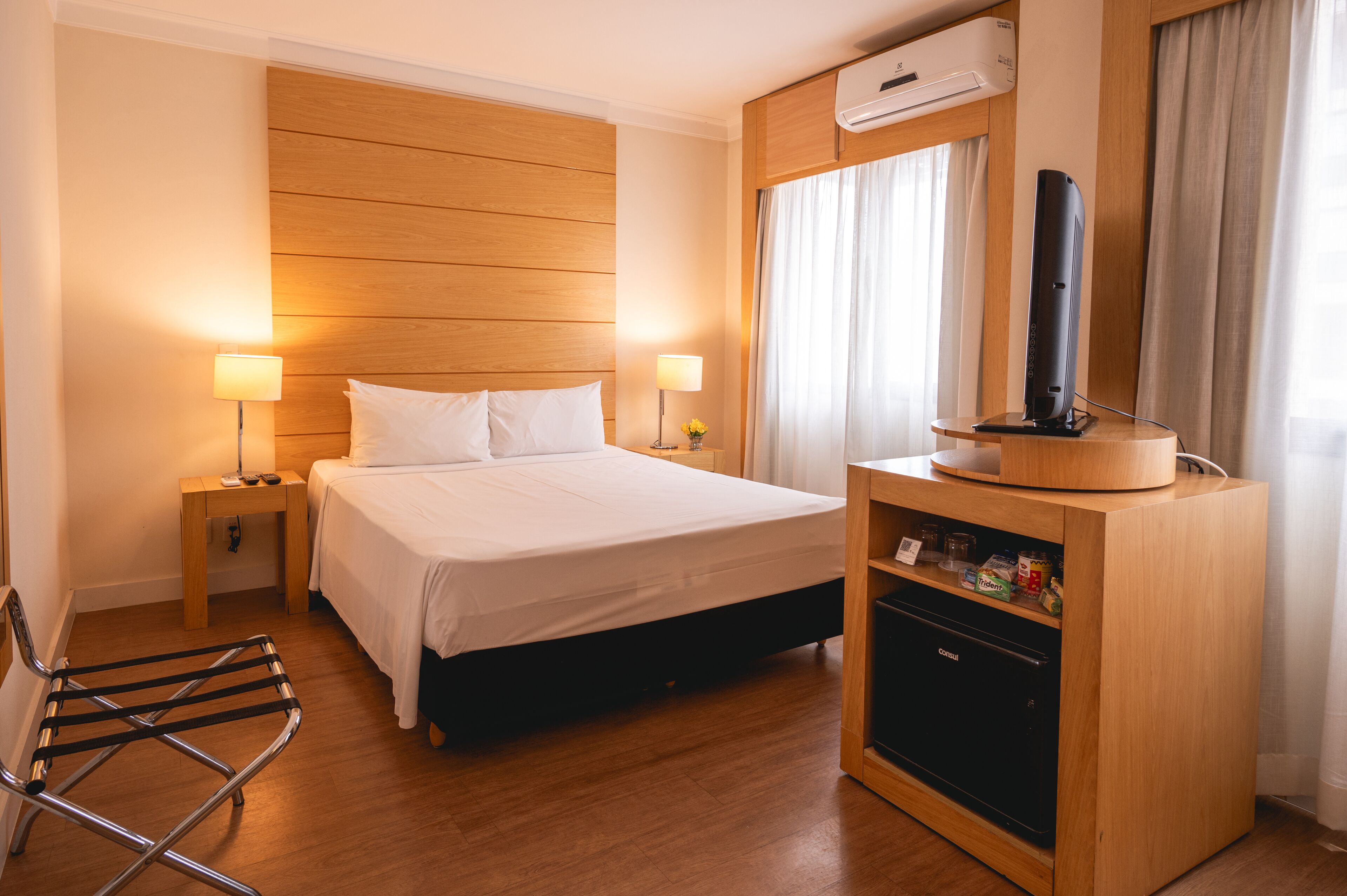 Habitación de hotel con cama, lámparas de noche, televisor en un mueble de madera y minibar.