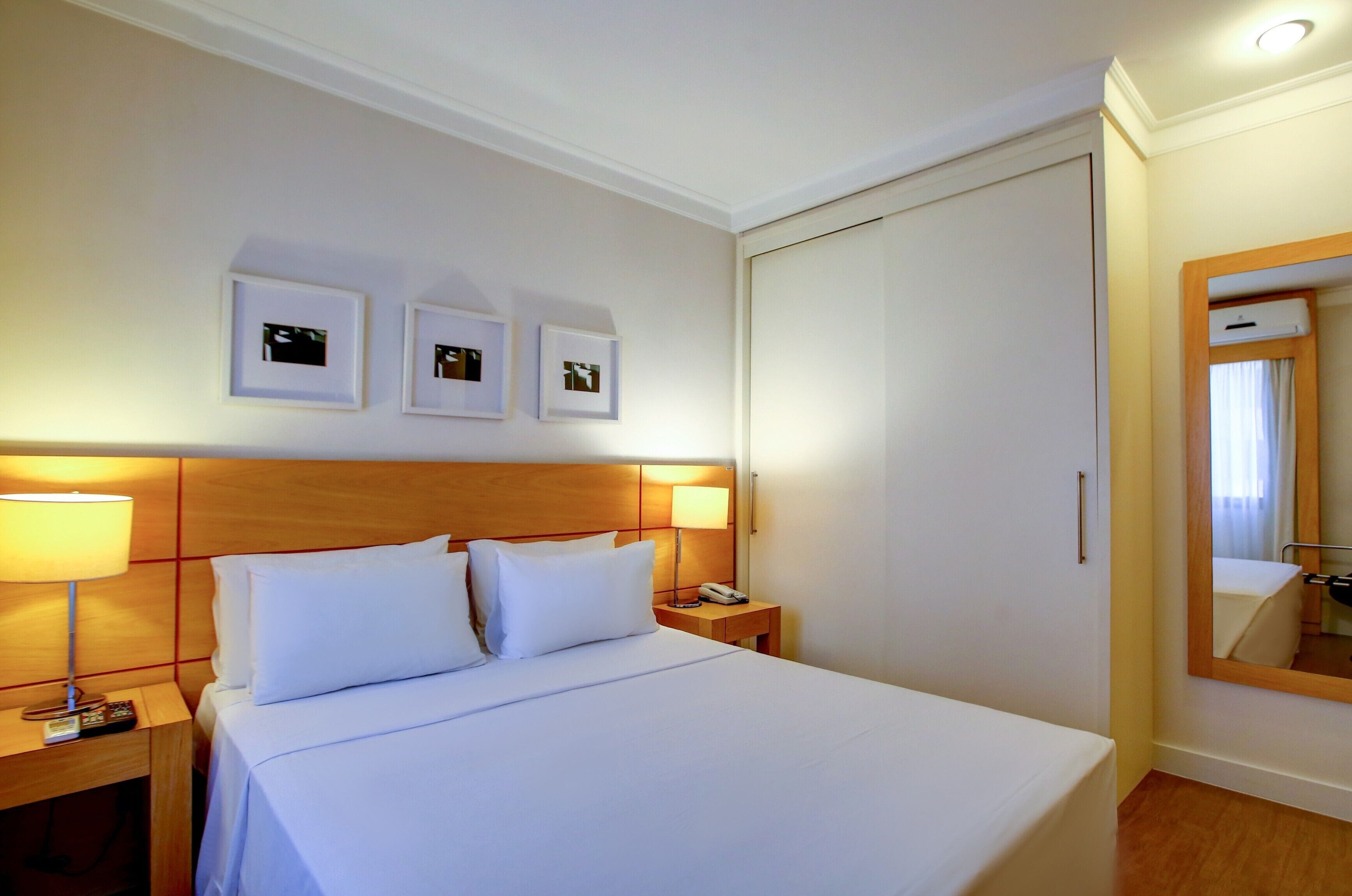 Una habitación de hotel con cama, lámparas de noche, mesita de noche, espejo y tres cuadros en la pared.