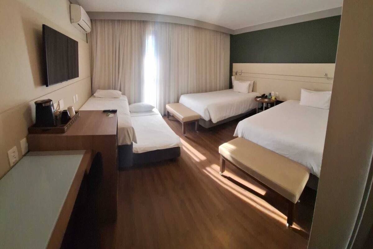 A hotel room with three single beds, a sofa bed, a desk with a chair, and a flat-screen TV mounted on the wall.